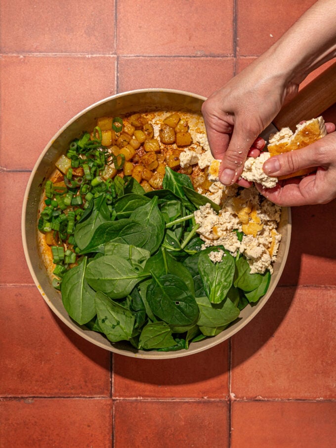 hand crumbling tofu onto skillet with spinach and potatoes