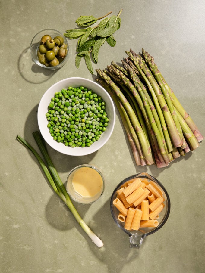 spears of asparagus, a white bowl with peas and a bowl of dry pasta