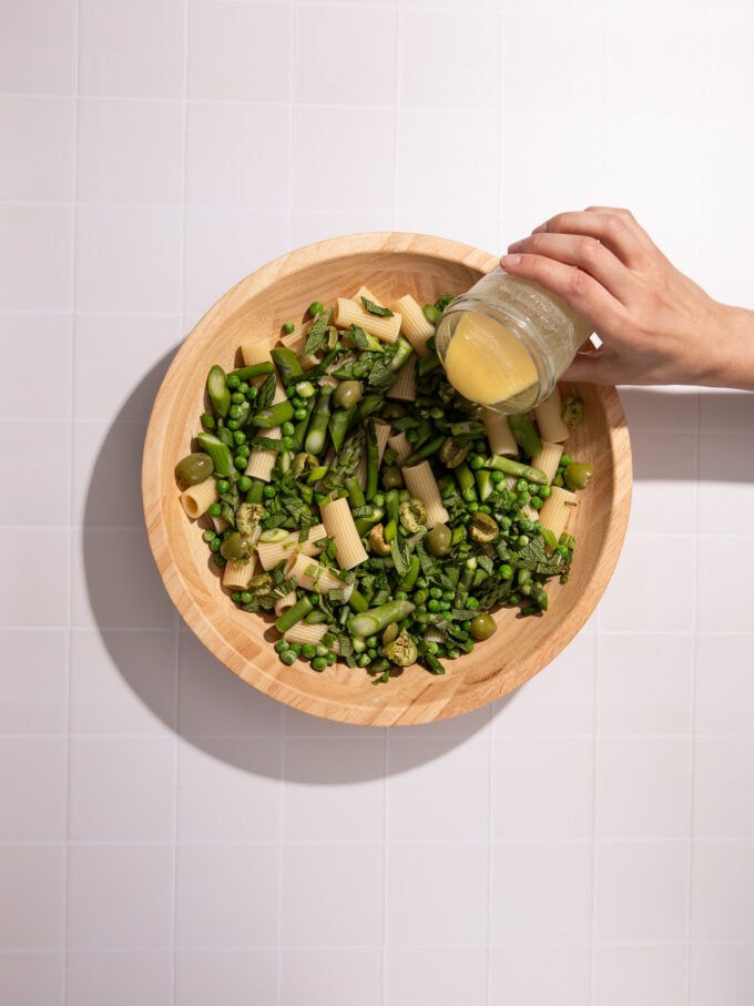 hand pouring dressing over asparagus and pasta in wood bowl.
