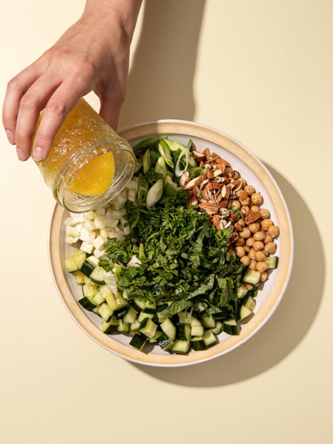 hand pouring dressing on salad bowl wiht chickpeas, cucumber and herbs