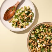 Chickpea cucumber salad on white plate with wooden spoon and bowl on yellow background