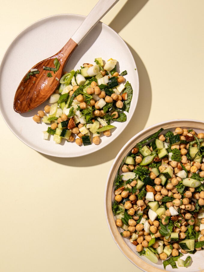 Chickpea cucumber salad on white plate with wooden spoon and bowl on yellow background
