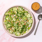 cucumber and peanut salad in a pink bowl with a jar of peanut sauce and spoon on the side