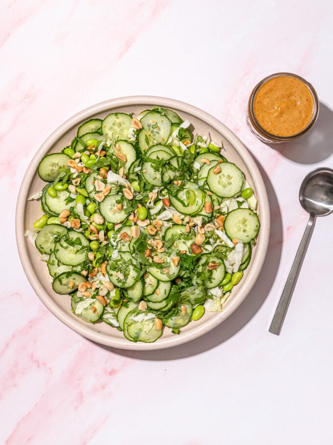 cucumber and peanut salad in a pink bowl with a jar of peanut sauce and spoon on the side