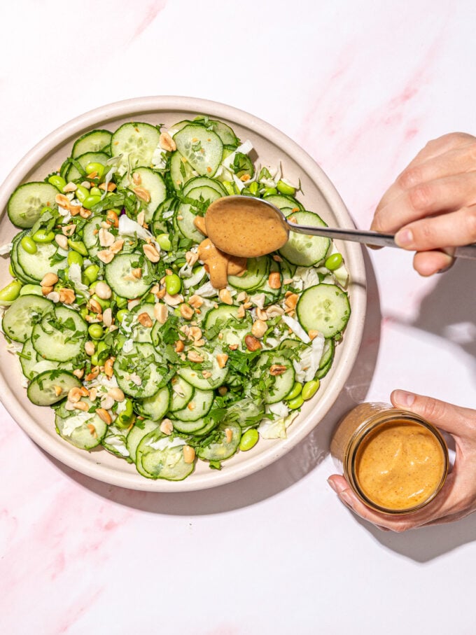 hand drizzling peanut sauce over a cucumber salad in pink bowl