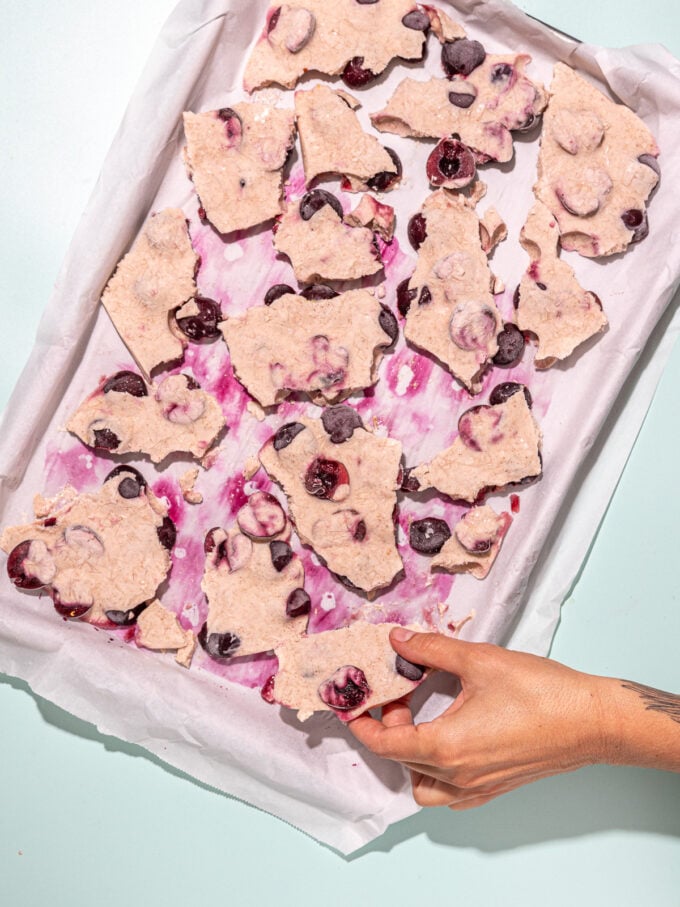 hand grabbing piece of cherry yogurt bark from baking sheet