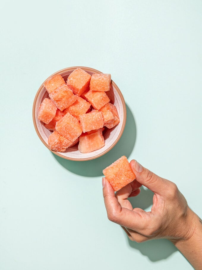 hand holding frozen watermelon chunk beside white bowl with watermelon
