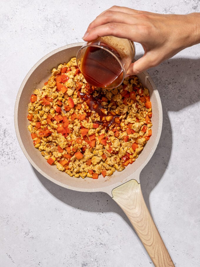 hand pouring sauce into skillet of tempeh and bell pepper