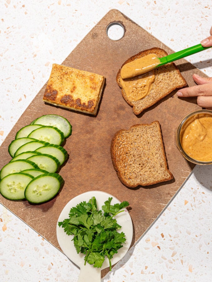 hand spreading peanut sauce on bread, with tofu, cucumber and cilantro on cutting board