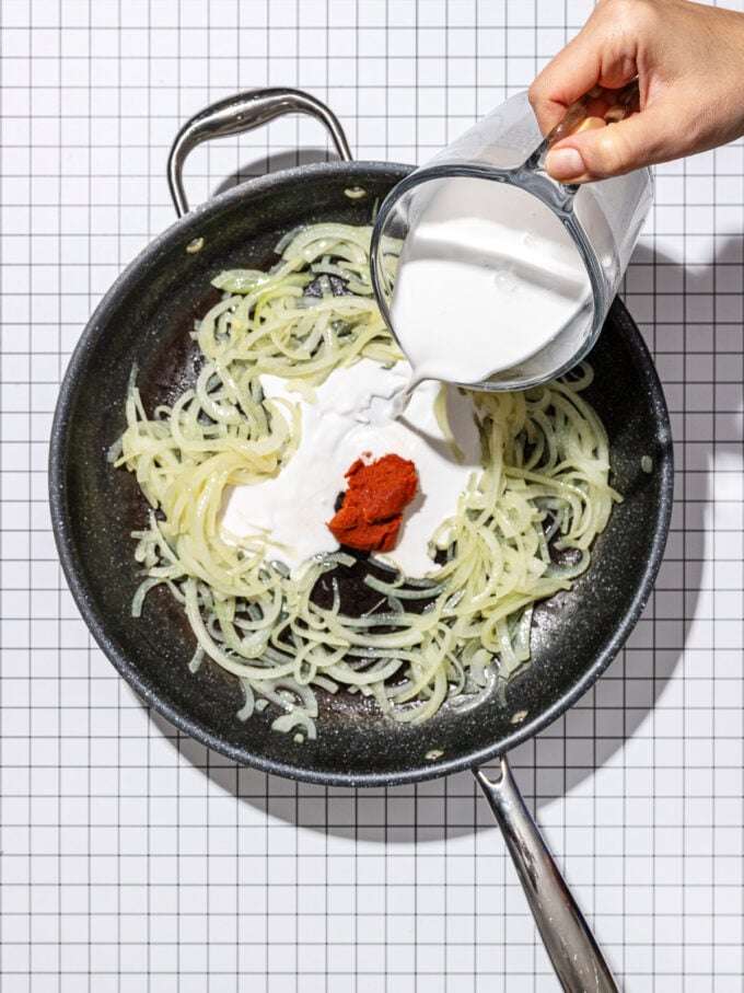 hand pouring coconut milk into pan of onions