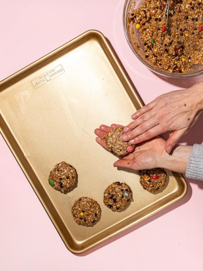 hands forming monster cookie with gold baking sheet and bowl of batter beside it