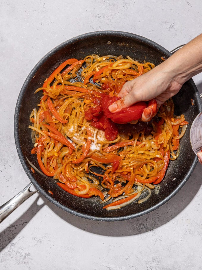hand crushing tomatoes into skillet with onion and peppers
