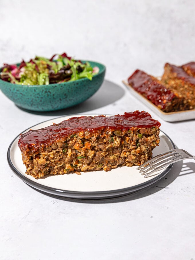 slice of lentil walnut loaf on white plate with blue bowl of salad behind it
