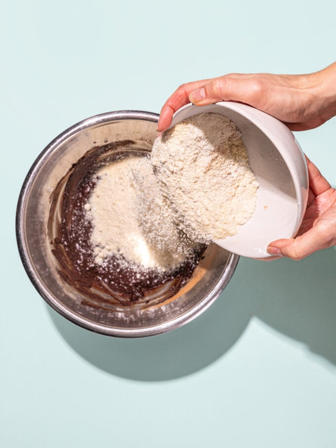 hand pouring almond flour into chocolate mixture in metal bowl