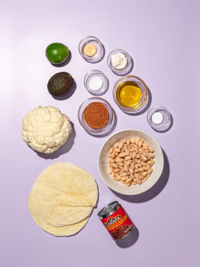 tortillas, can of chipotle peppers, head of cauliflower, bowl of white beans and spices on purple background