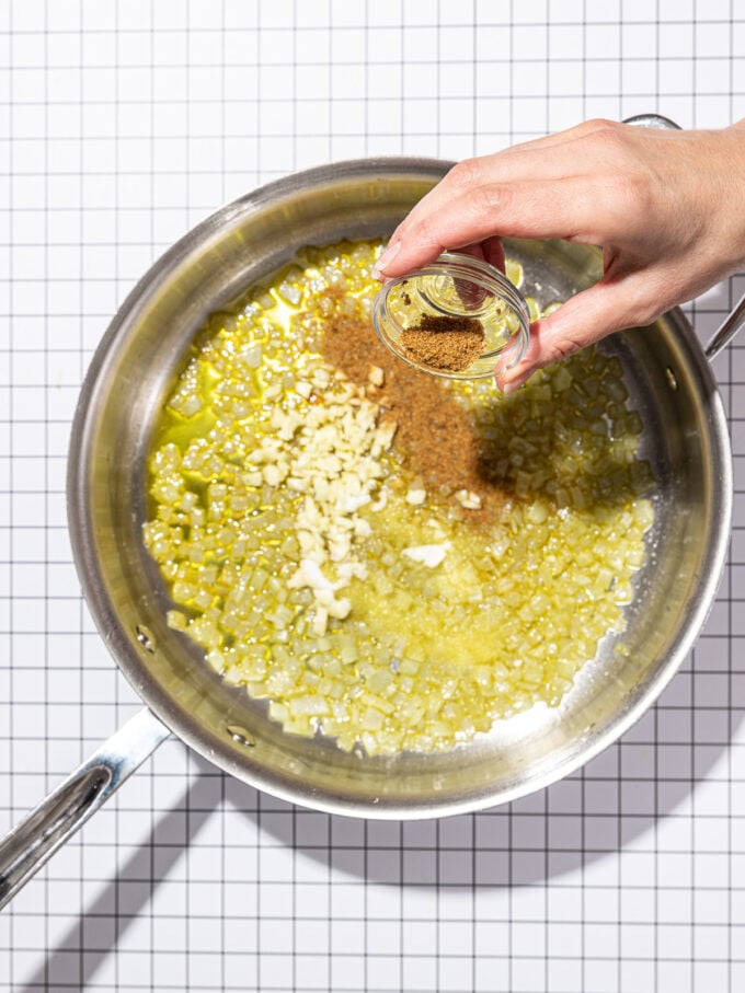 hand pouring cumin and garlic into cooked onions in skillet