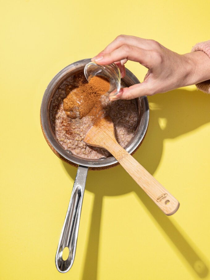hand pouring cinnamon into buckwheat porridge in metal pot