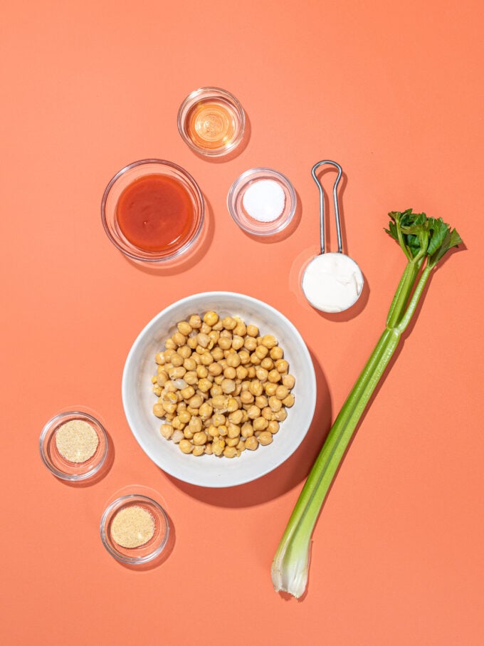 celery rib, bowls of chickpeas, buffalo hot sauce, mayo and salt on orange background