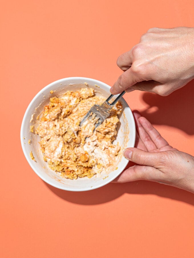hand mixing smashed chickpeas and mayo in white bowl with fork
